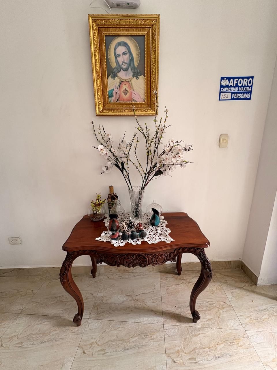 A wooden table with a lace doily, a framed religious painting, and a sign indicating room capacity.