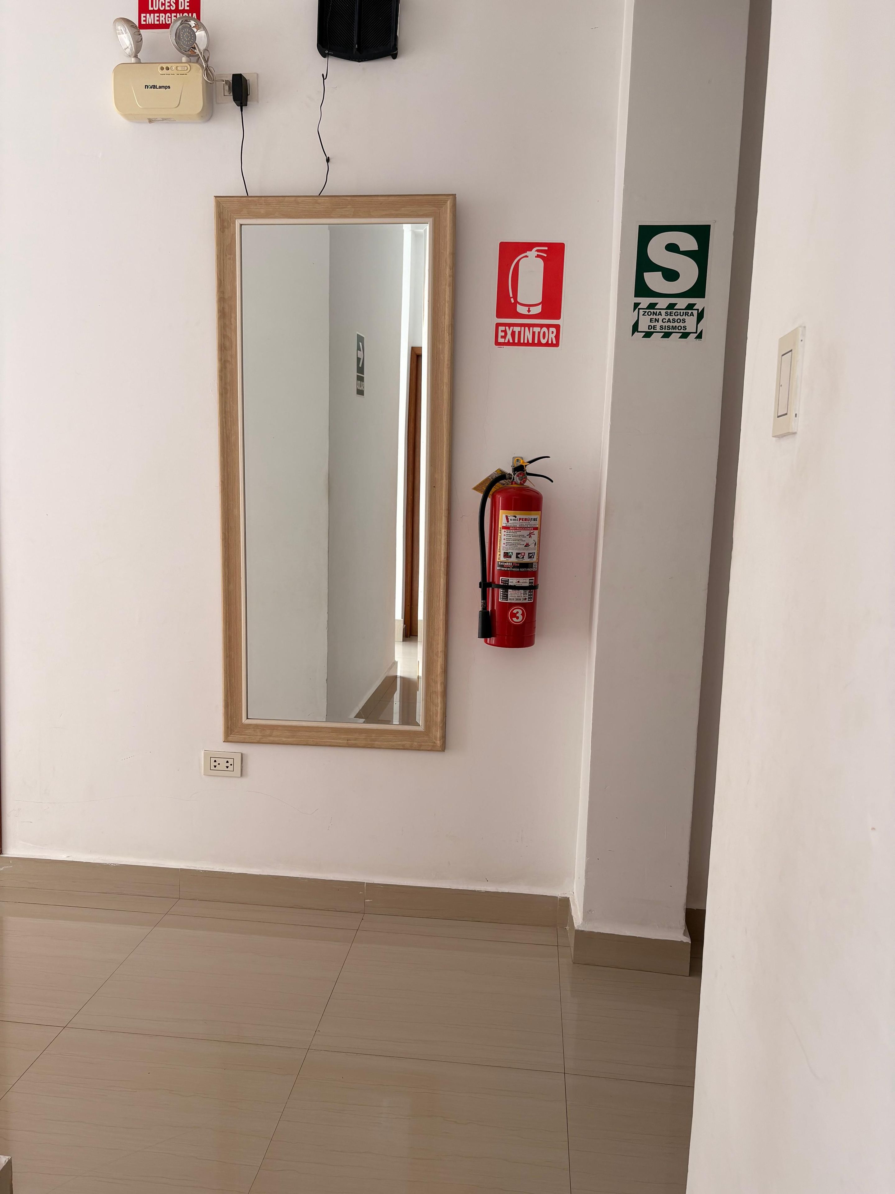 A hallway with a mirror, a fire extinguisher, a security zone sign, and a light switch.
