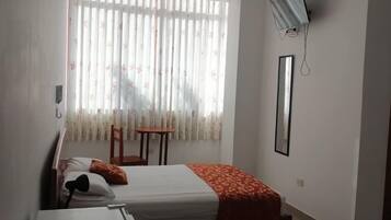 A hotel room with a bed, a small table, a chair, a TV mounted on the wall, and a window with curtains.