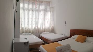 Una habitación de hotel con dos camas individuales, cabecero de madera, una mesita de noche pequeña, teléfono en la pared y ventana con cortinas florales.