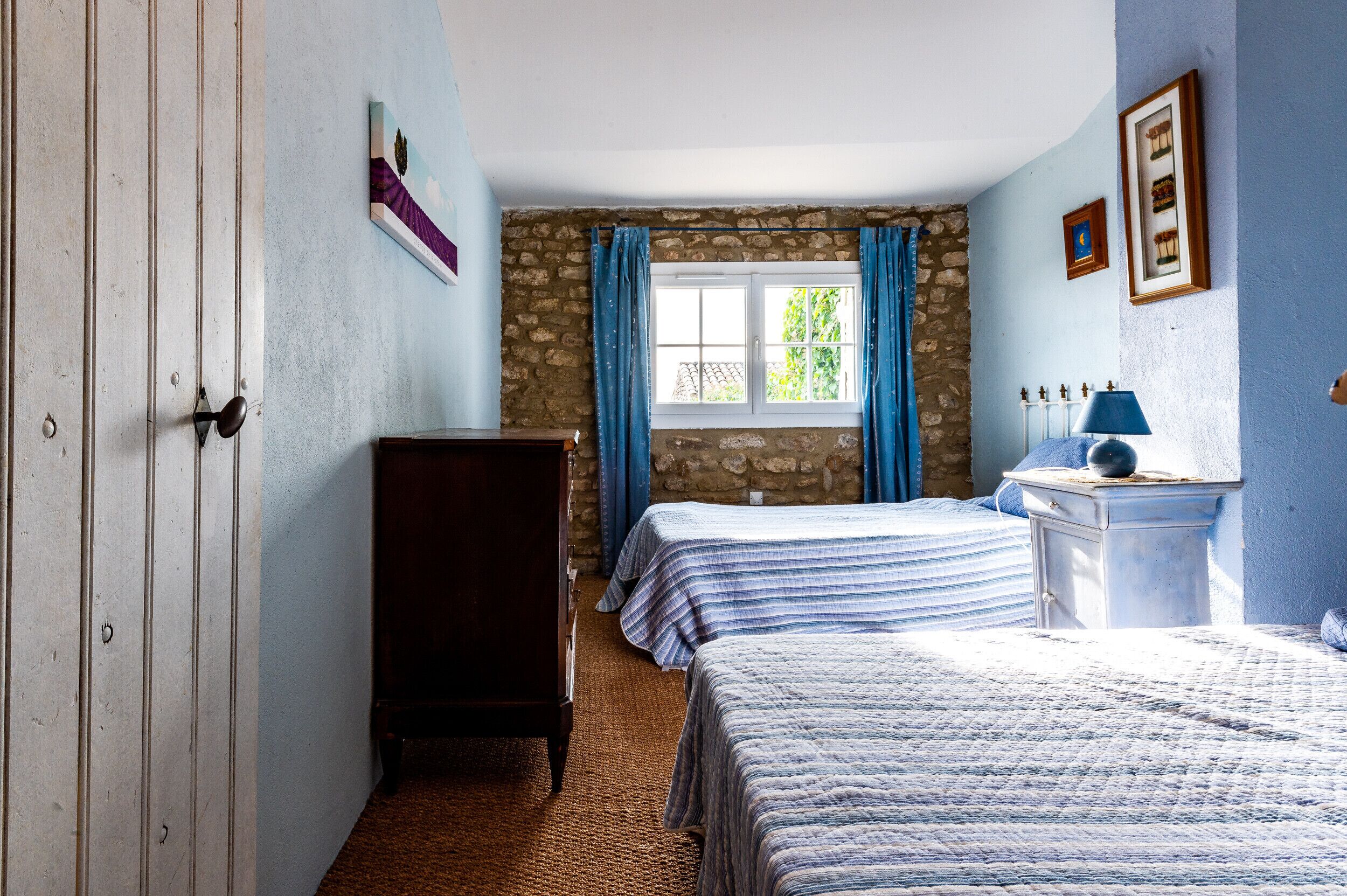 Ein Zimmer mit zwei Betten, einem Holzschrank, einer Steinsäule, einem Fenster mit blauen Vorhängen und einem Nachttisch mit Lampe.