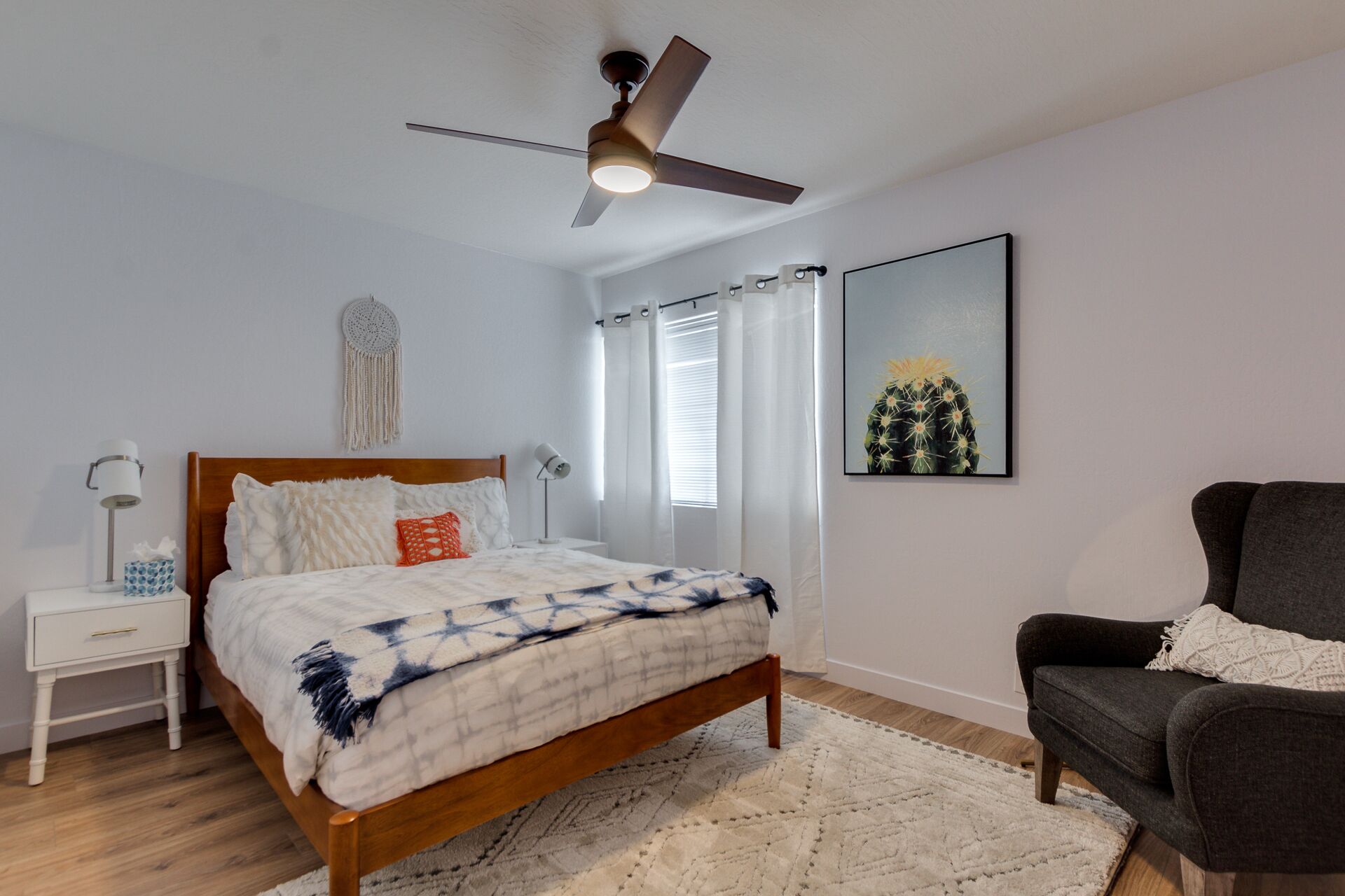Une chambre à coucher avec un lit en bois, une table de chevet, un bureau, une chaise, un ventilateur de plafond et une fenêtre avec des rideaux.