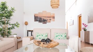A modern living room with a glass coffee table, a beige sofa with green patterned cushions, a large potted plant, and a wall-mounted artwork depicting a beach scene. - Unique Family Villa! Spa, Ocean view, Heated Pool, Rooftop (Antigua)