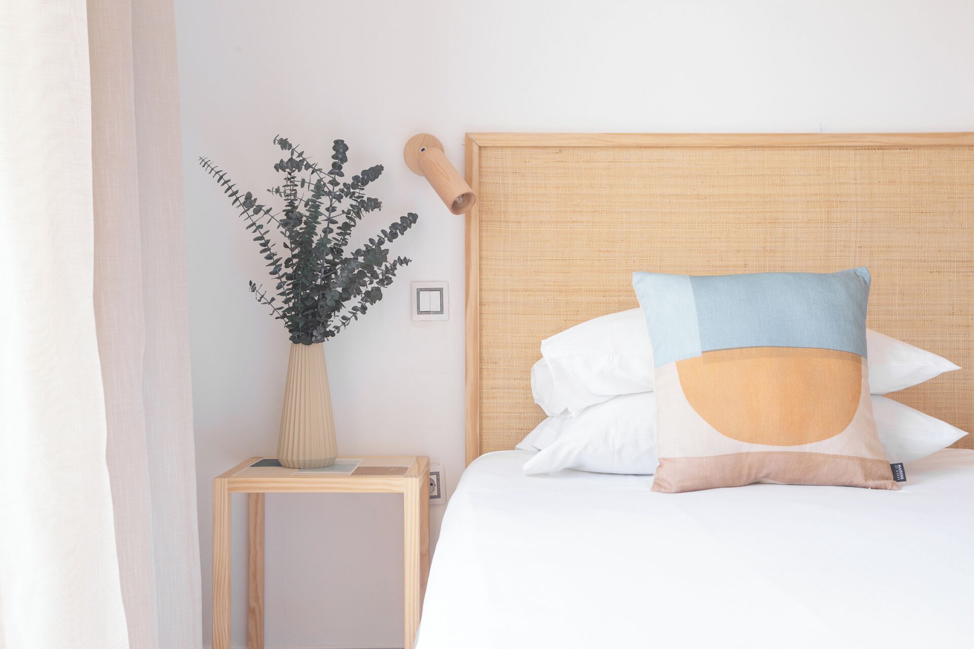 Una cama con cabecera, una mesita de noche con un florero y un interruptor de pared.
