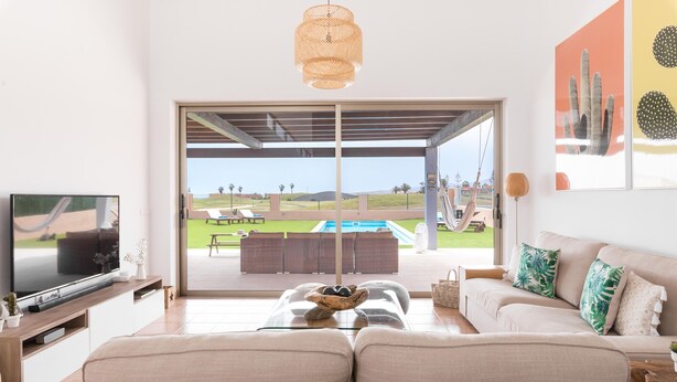A spacious living room with a large flat-screen TV, a glass coffee table, and a beige sofa. There is a large sliding glass door leading to an outdoor patio with a pool and lounge chairs. - Unique Family Villa! Spa, Ocean view, Heated Pool, Rooftop (Antigua)
