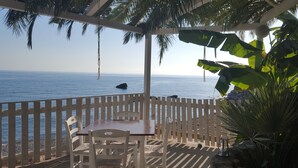 Terraza de madera con una mesa y sillas, con vista a un mar tranquilo y un promontorio rocoso.