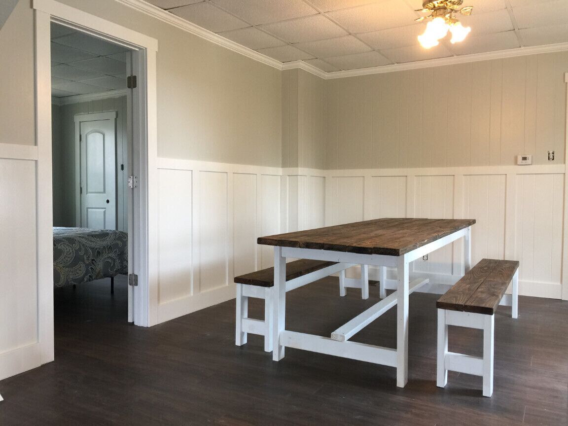 A room with a wooden dining table and benches, white paneled walls, and a dark wooden floor.