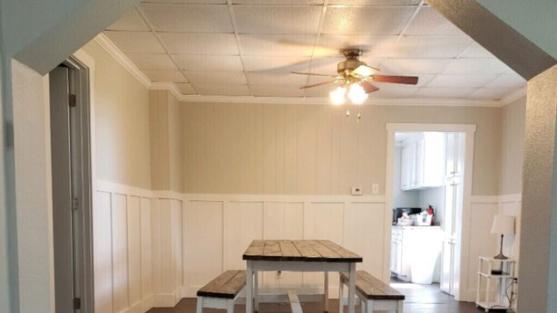 Un espace repas avec une table et des bancs en bois, des murs recouverts de panneaux blancs et un ventilateur de plafond.