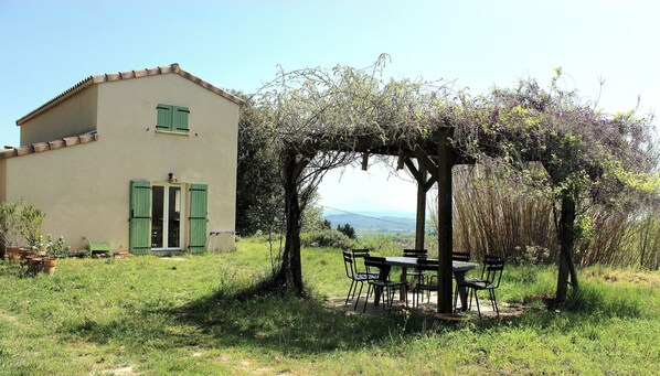 A house with a garden, a pergola with a table and chairs, and a view of the landscape.