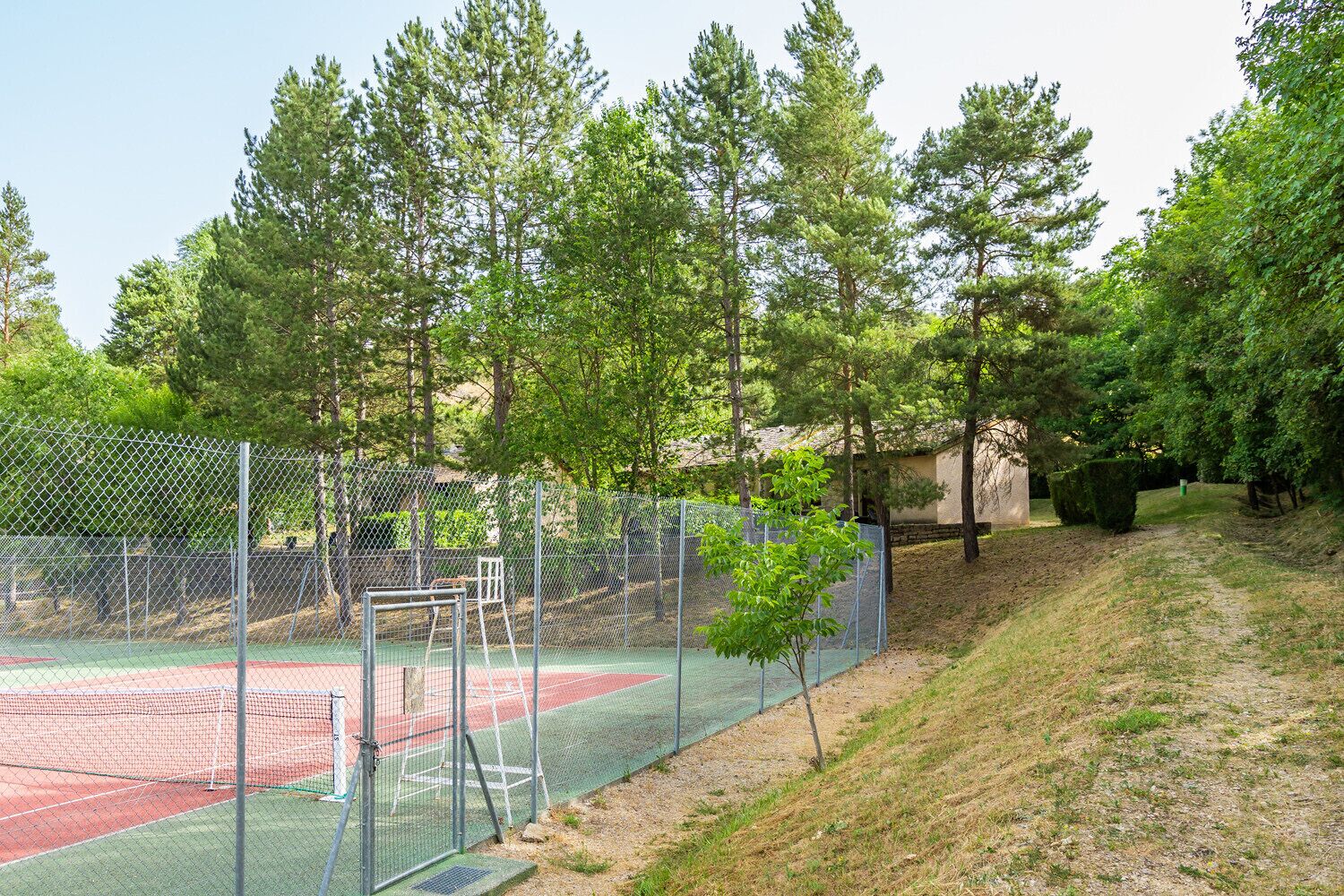 A tennis court surrounded by a chain-link fence, with trees and a building in the background.