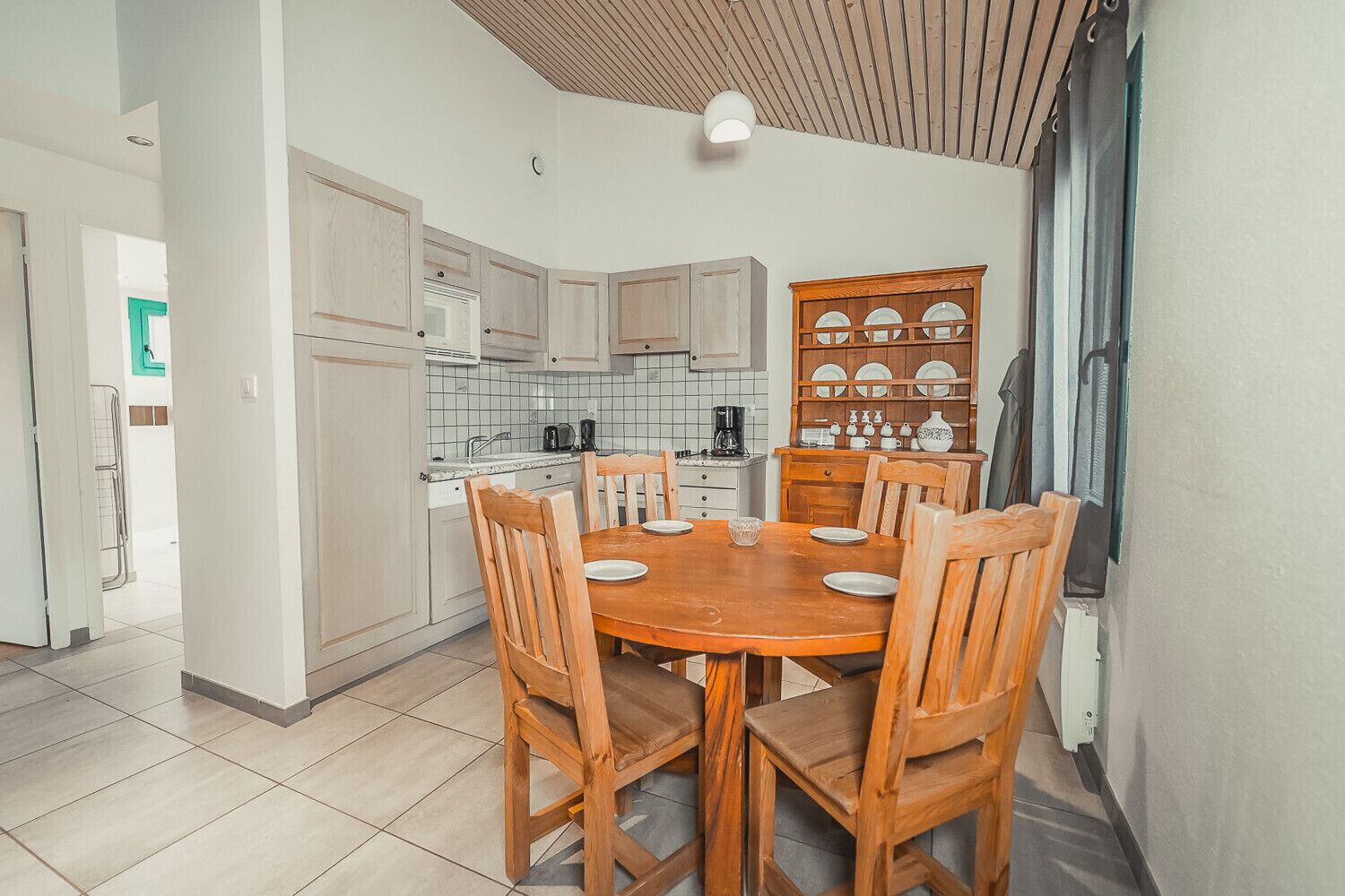 Cocina compacta con armarios blancos, una mesa de madera con sillas y una cafetera.