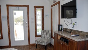 A room with a wooden door, a patterned chair, a TV mounted on the wall, and a cabinet with electronic devices. - White Oak -Beautiful Cottage - Perfect For a  Family Getaway (Aransas Pass)