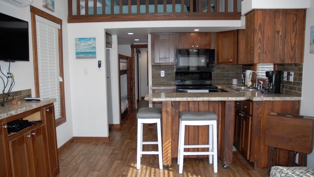 A kitchen with wooden cabinets, a central island with stools, a built-in oven, and a television mounted on the wall. - White Oak -Beautiful Cottage - Perfect For a  Family Getaway (Aransas Pass)