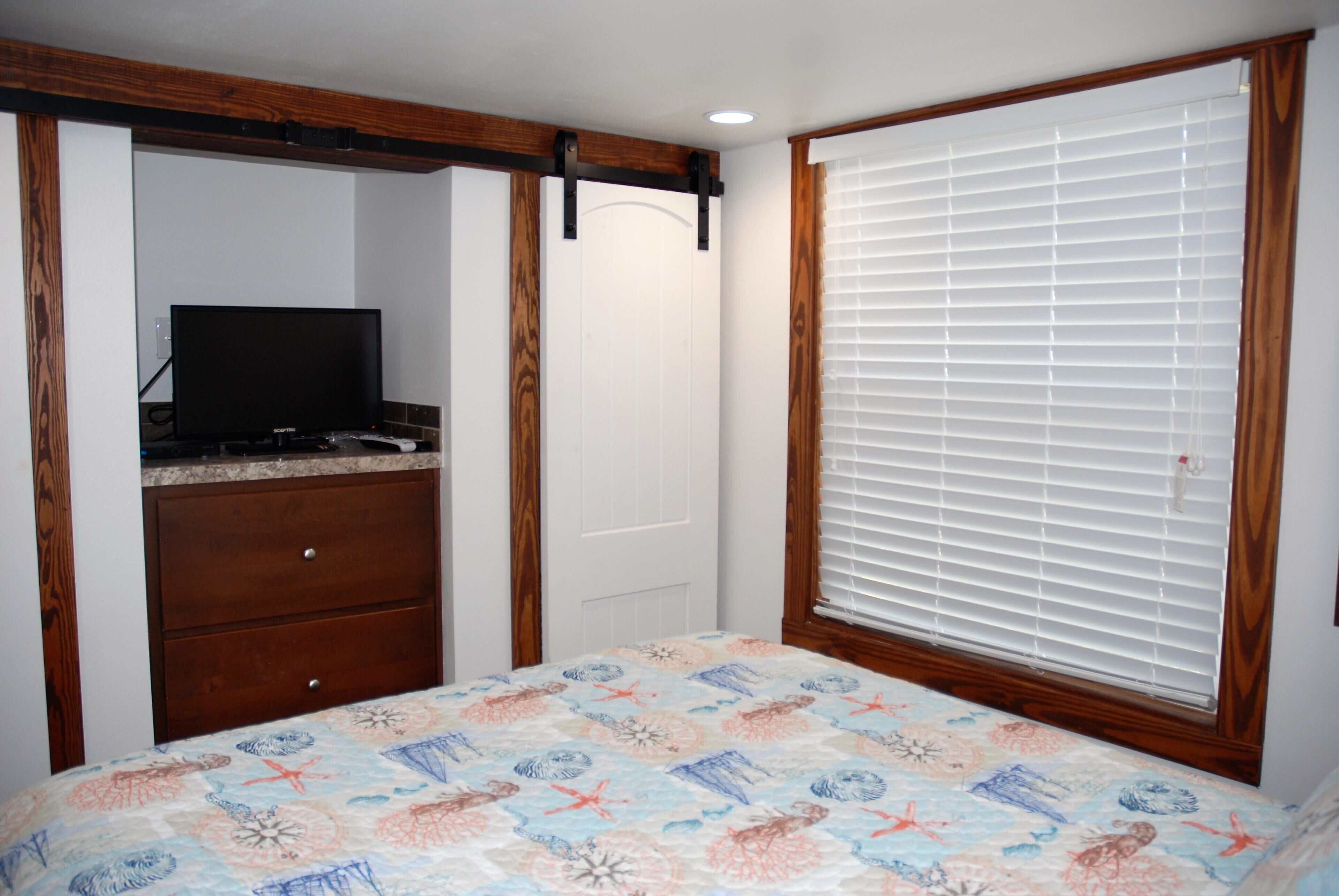 Une chambre à coucher comprenant un lit, une commode, un téléviseur et une fenêtre avec des stores.