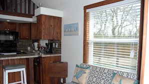 A kitchen with wooden cabinets, a built-in microwave, and a bar stool. There is a window with blinds and a sofa with patterned cushions. - White Oak -Beautiful Cottage - Perfect For a  Family Getaway (Aransas Pass)