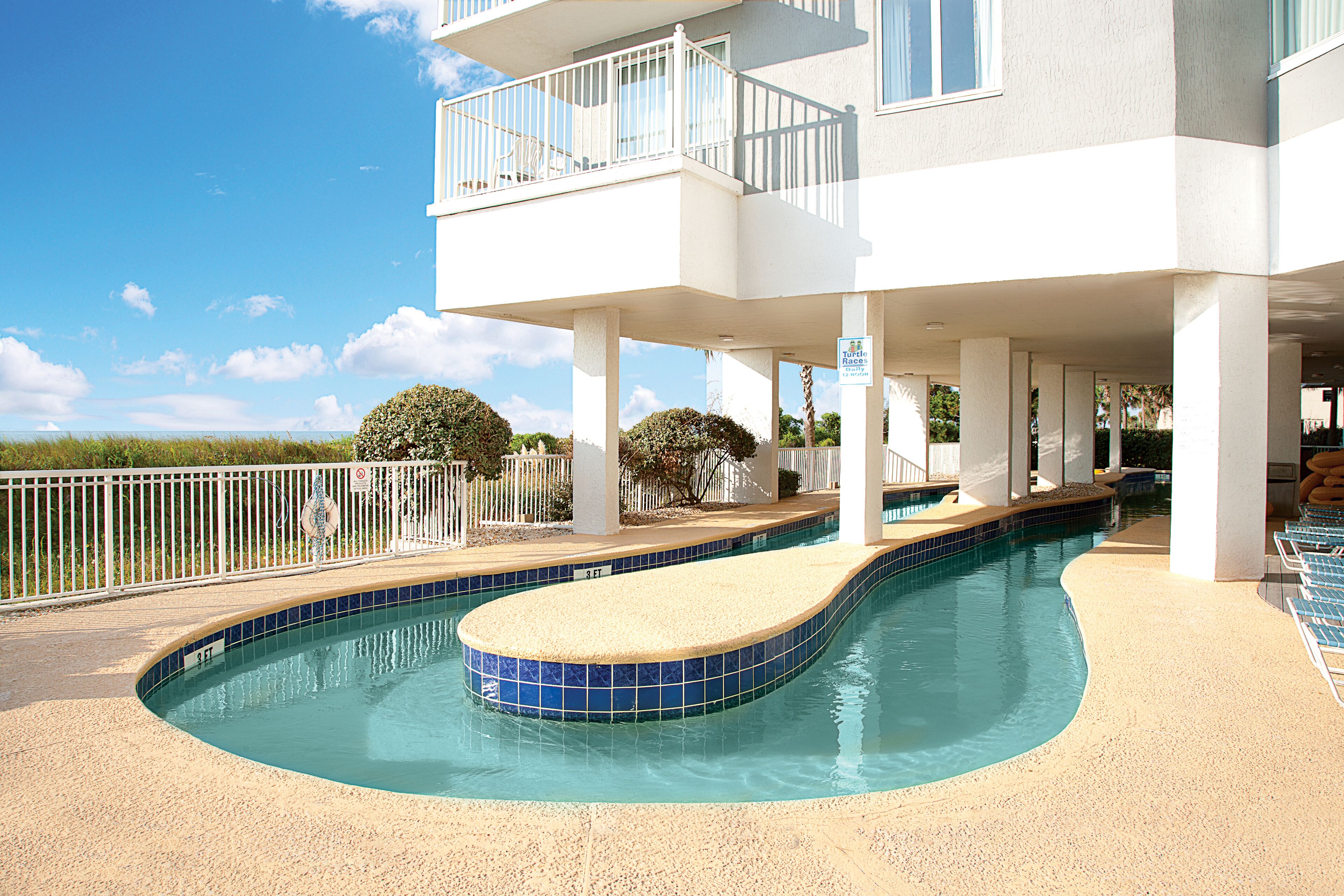 A swimming pool with a curved edge, a white building with balconies, and a clear blue sky.