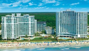 Un complejo hotelero frente a la playa con varias torres altas, una zona de piscina y una playa con sombrillas y gente.
