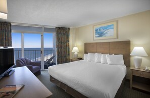 Habitación de hotel con una cama grande, dos lámparas de noche, un escritorio con silla y vista al océano a través de puertas corredizas de vidrio.