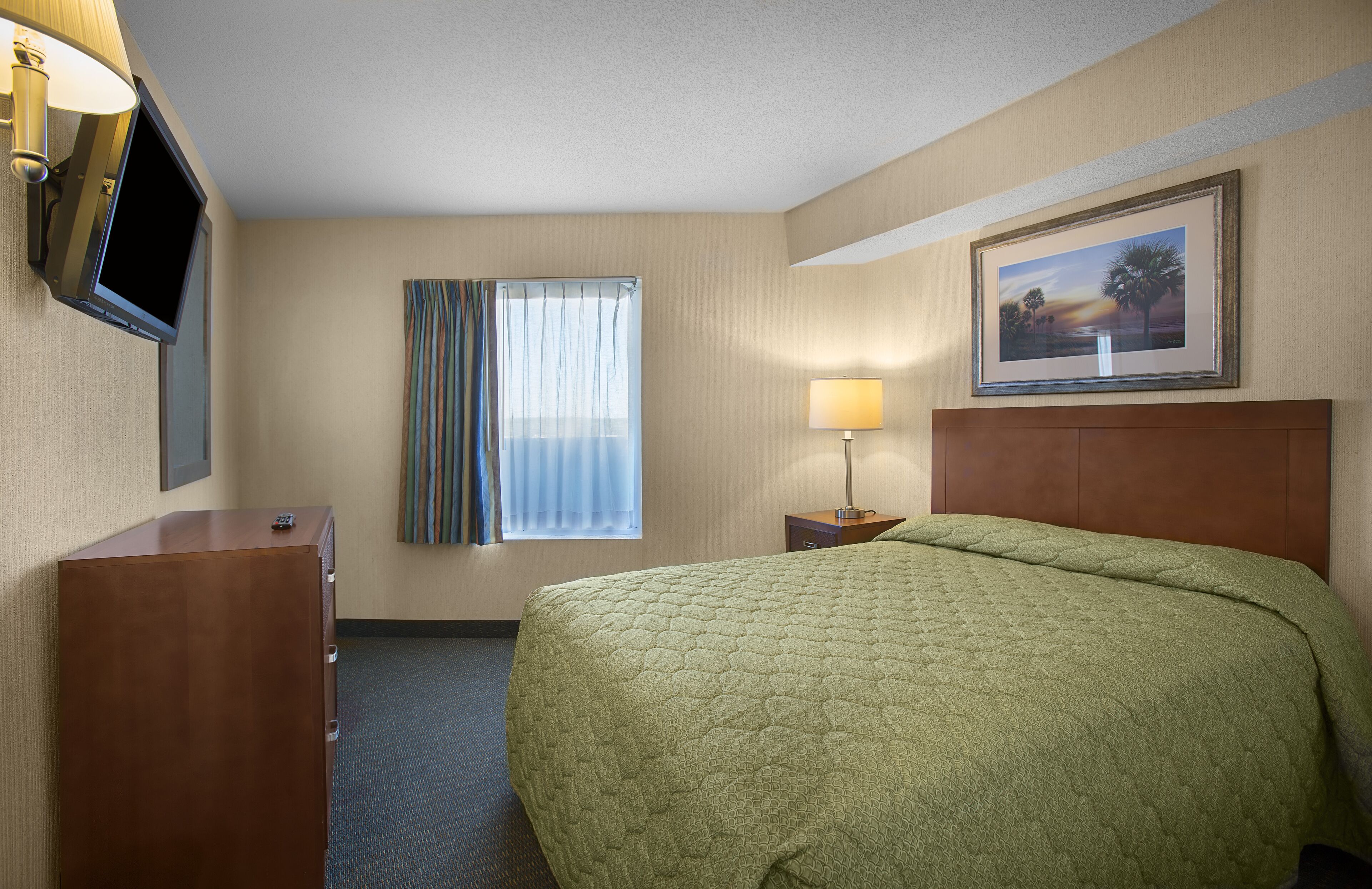 A hotel room with a bed, a TV mounted on the wall, a window with curtains, a nightstand with a lamp, and a framed picture on the wall.