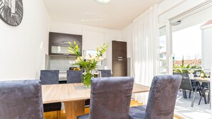 Un espace repas moderne avec une table en bois et des chaises à revêtement sombre, une cuisine en arrière-plan et une vue sur l’extérieur grâce à de grandes fenêtres.