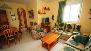 Ein Wohnzimmer mit einer Couch, Sesseln, einem Couchtisch, einem Fernseher und einem Esstisch.