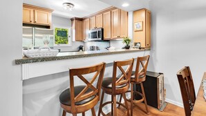 Cocina moderna con armarios de madera, un refrigerador de acero inoxidable, un microondas y una encimera de granito. Hay dos taburetes de bar de madera y un refrigerador pequeño.