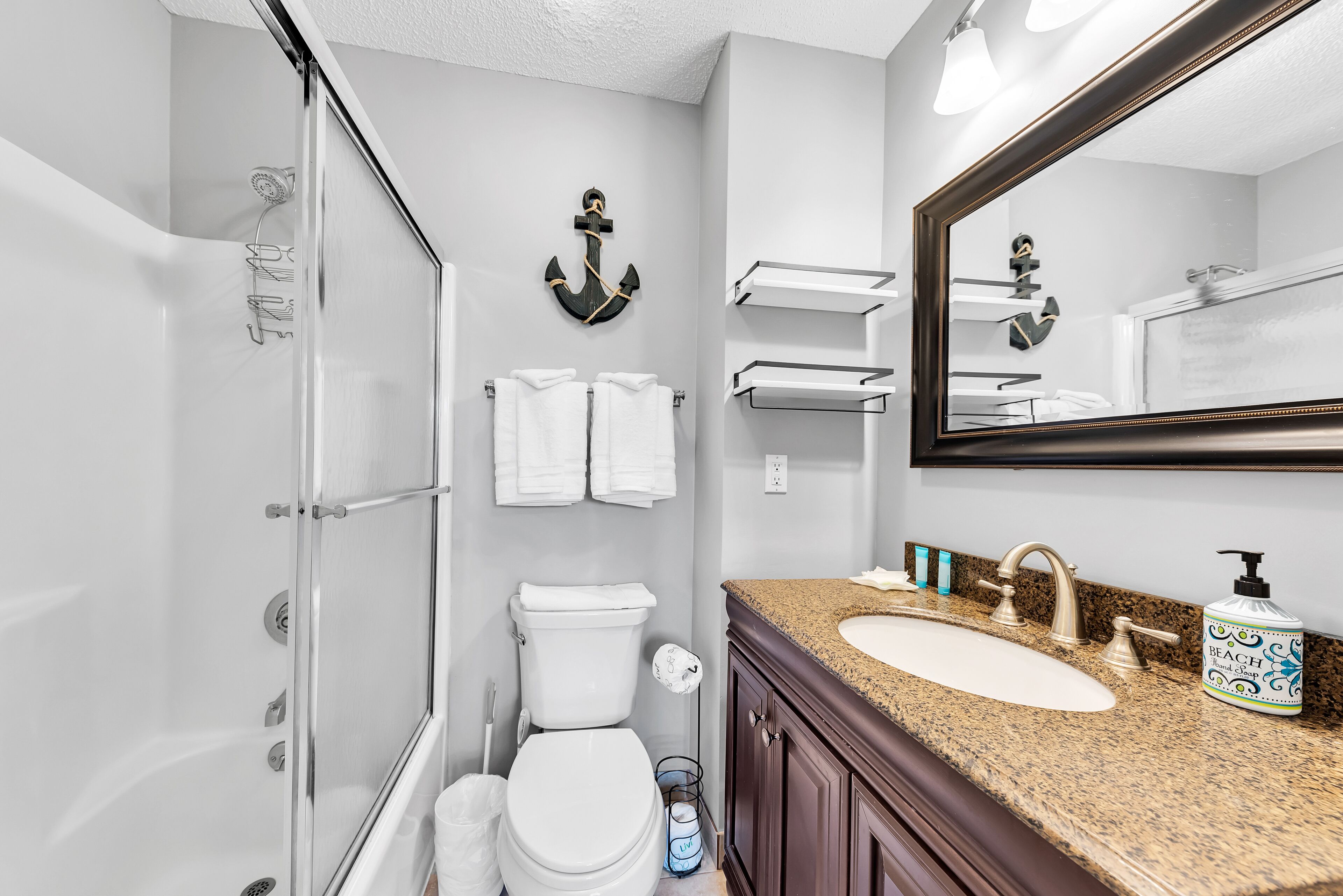 A bathroom with a glass shower enclosure, a toilet, a sink with a granite countertop, and a mirror with an anchor decoration.