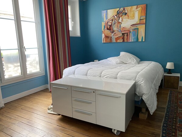 A bedroom with a bed, a bedside table, a lamp, a painting on the wall, and a window with curtains. - PORTE MAILLOT & ETOILE - IDEAL LOCATION FOR WORK AND TOURISM - LUXURIOUSLY RENOVATED APARTMENT (Paris-16)