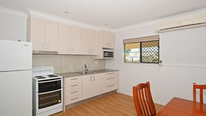 A compact kitchen with light wood cabinets, a white refrigerator, a stove, and a microwave. There's a window with a view of the outside, and a wooden dining table with chairs. - Hami's Retreat - Burrum Heads- Close to Boatramp -2BR - Aircon (Burrum Heads)
