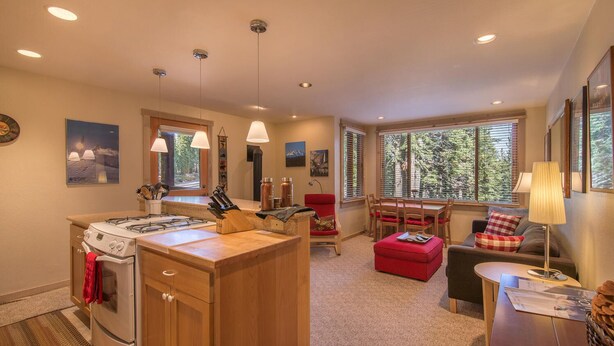 Una cocina moderna con una isla central, comedor con una silla roja y una zona de estar acogedora con una lámpara.