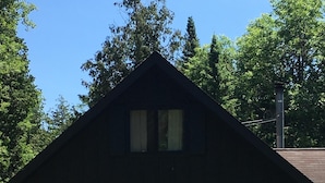 Una cabaña de madera de dos pisos con un porche y una silla, rodeada de follaje y árboles.