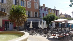 An outdoor café with tables and chairs, a fountain, and colorful buildings in the background. - Apartment in the heart of the village • 50 m² terrace (Saint-Paul-Trois-Châteaux)