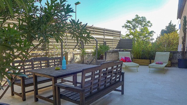 Un patio al aire libre con una mesa y bancos de madera, rodeado de plantas en macetas y una cerca de metal.