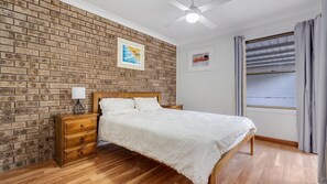 A bedroom with a brick-patterned wall, a wooden bed, a nightstand, a ceiling fan, and a window with curtains.