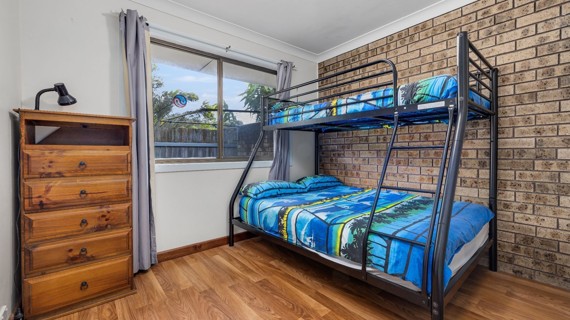 A bunk bed with a blue and yellow patterned bedspread, a wooden dresser with four drawers, a window with curtains, and a brick wall.