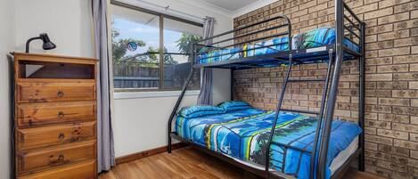 A bunk bed with a blue and yellow patterned bedspread, a wooden dresser with four drawers, a window with curtains, and a brick wall.