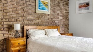A bedroom with a brick wall, a wooden bed, a nightstand with a lamp, and two framed pictures. - Cresta 1 Crescent Head (Crescent Head)