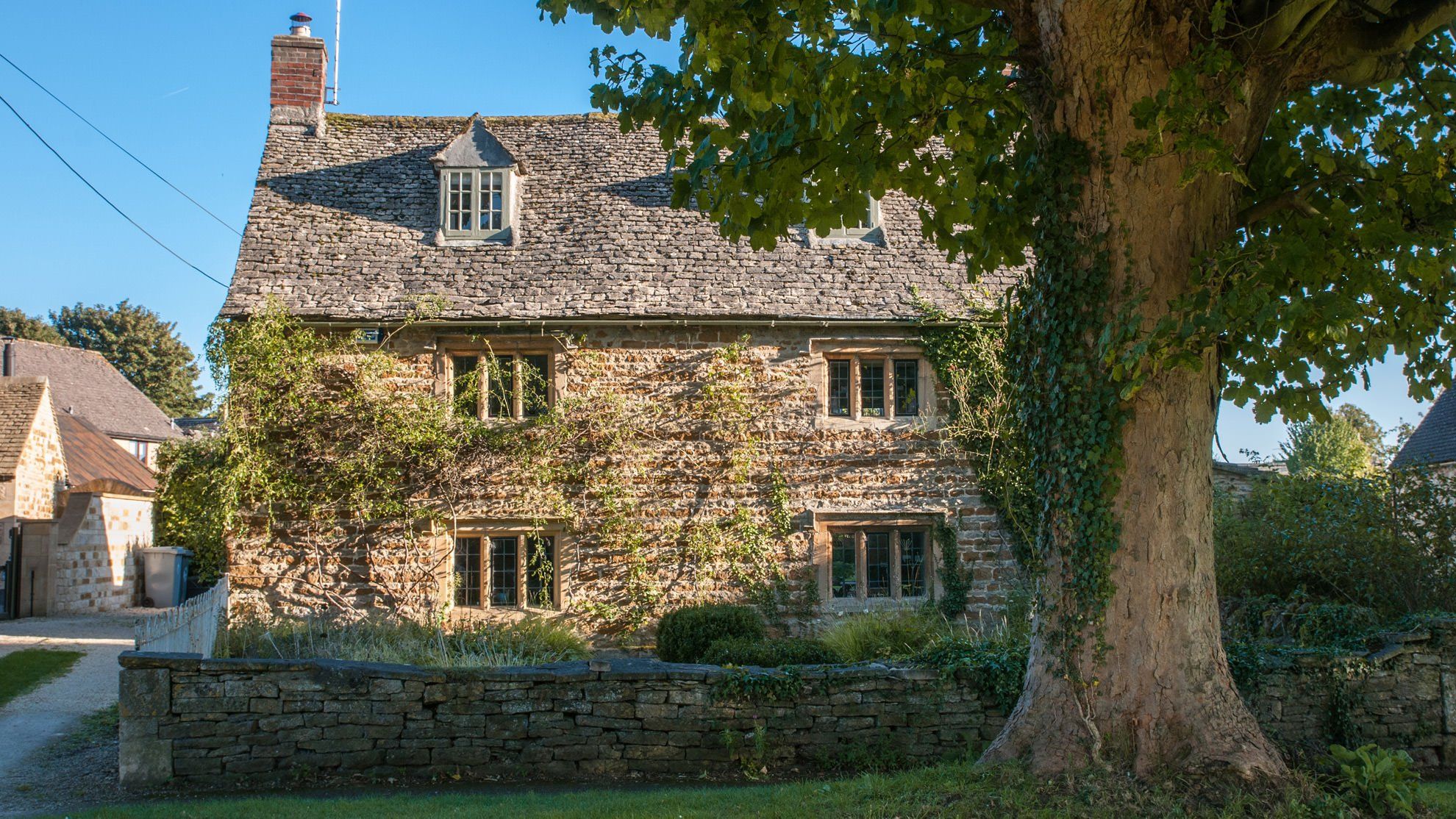 A stone house with multiple windows and a chimney, surrounded by greenery and a stone wall.