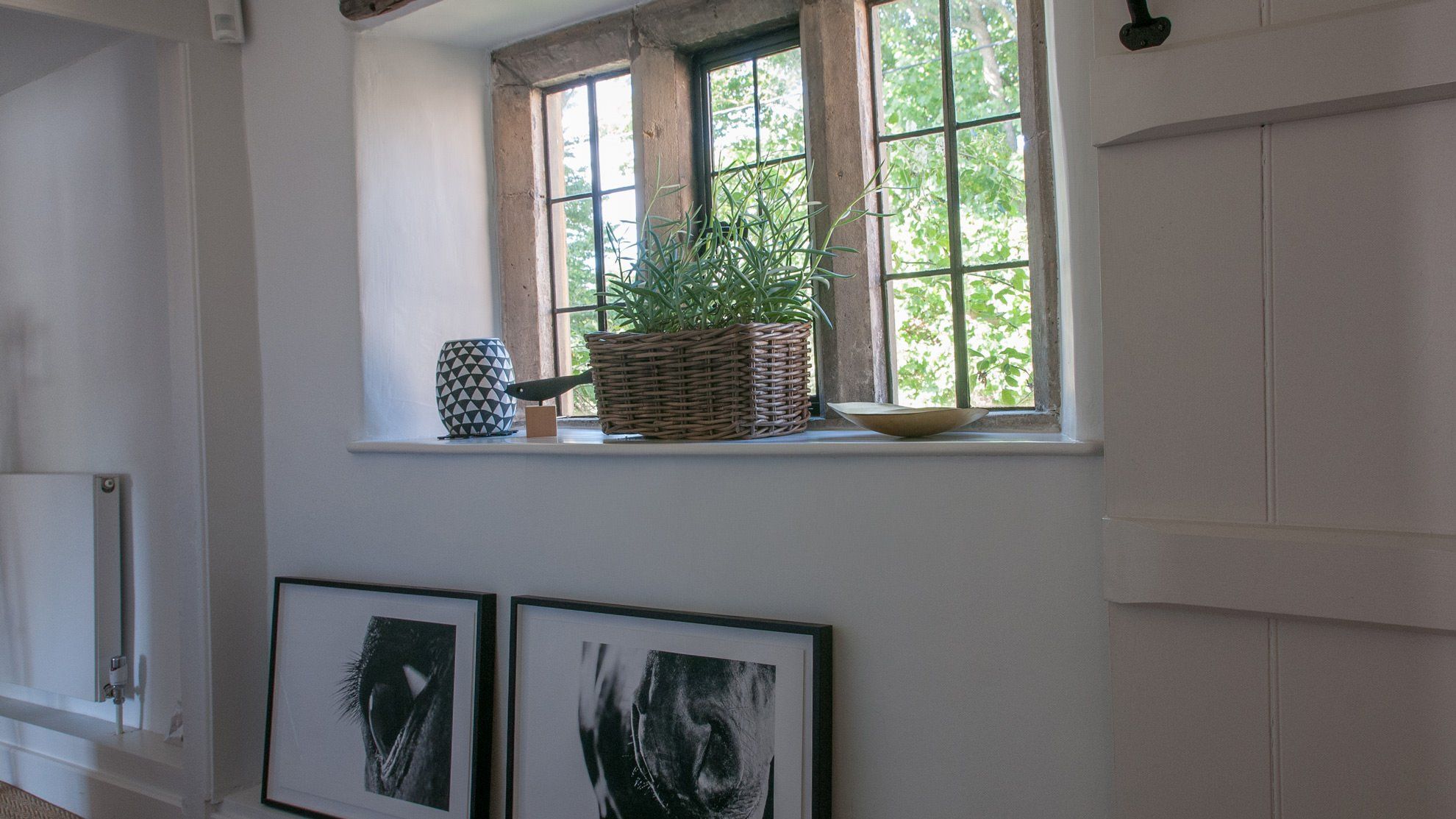 Una habitación con ventana, una cesta de mimbre con plantas, un florero estampado y dos cuadros en la pared.