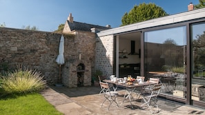 A stone wall, a brick chimney, a stone oven, a glass door leading to a kitchen, and a table with chairs outside. - Bentleys - Three Bedroom Cottage, Sleeps 6 (Kingham)