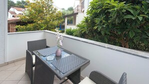 Un balcon agrémenté d’une table en verre et de chaises en osier, entouré de verdure et bordé d’un mur blanc.