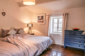 Une chambre à coucher avec un lit, une commode, une lampe, un tableau au mur et une fenêtre avec des rideaux.