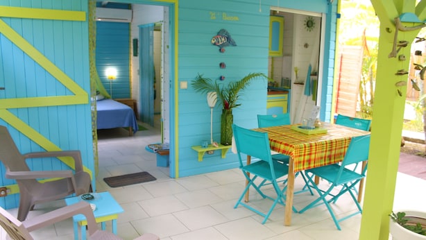 A room with a bed, a small table with chairs, a window, and a door leading to another room. - Coco Bungalows. Bungalows 2 / pers sea at 100m (Ste Rose)
