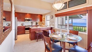 A modern kitchen with a central island, dining table with chairs, and a view of the ocean through large windows. - Kapalua Ridge Villa 1124 Luxury Oceanview Retreat (Lahaina)