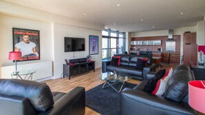 A modern living room with a black leather sofa, a glass coffee table, and a flat-screen TV. A kitchen with a stainless steel range hood is visible in the background. - Lancefield Quay - SECC & Hydro Arena, parking available (Glasgow)