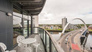 A balcony with white metal chairs and tables overlooking a cityscape, a river, and a bridge.