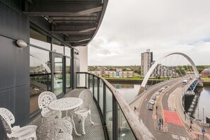 Un balcon avec des chaises et des tables en métal blanc, d’où l’on peut admirer un paysage urbain, une rivière et un pont.