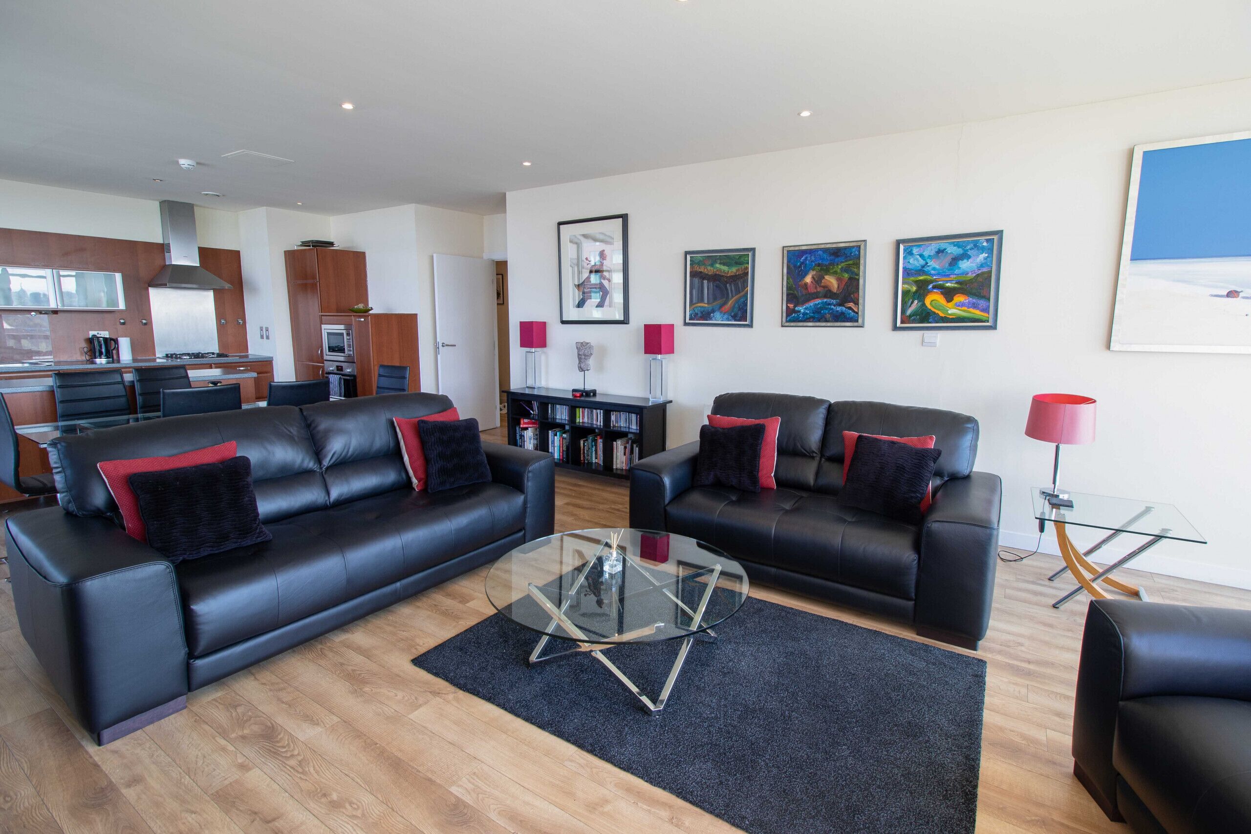 A modern living room with two black leather sofas, a glass coffee table, and a kitchen in the background.