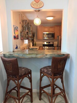 A kitchen with wooden cabinets, a stainless steel refrigerator, and a built-in oven. There are two wicker chairs with floral upholstery at the kitchen island.