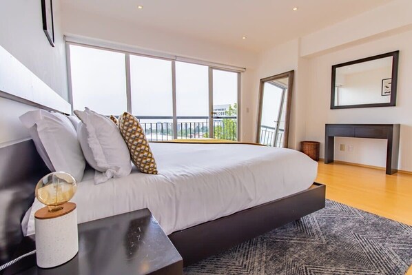 Une chambre à coucher moderne avec un grand lit, une table de chevet, un miroir et une vue sur l’extérieur.